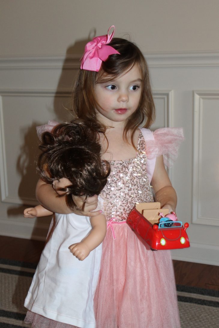 Little girl with pink hair bow and pink glitter dress carrying an American Girl Doll