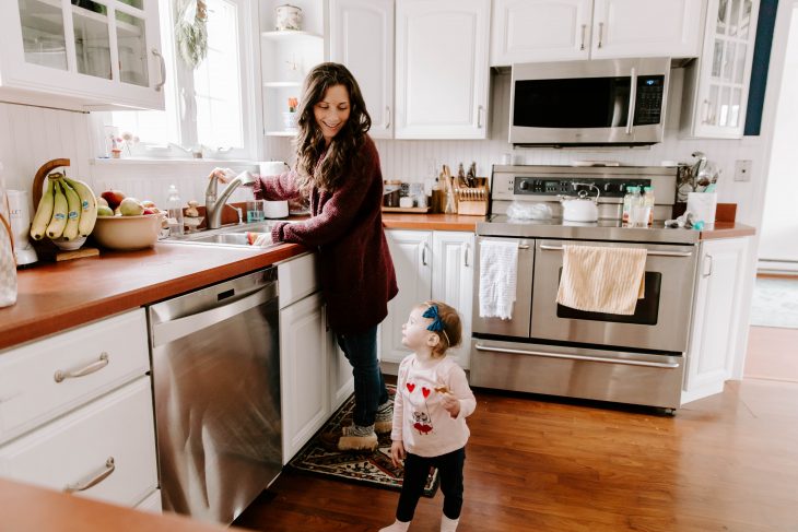 Woman cleaning dishes in the kitchen while toddler looks up at her to simplify life