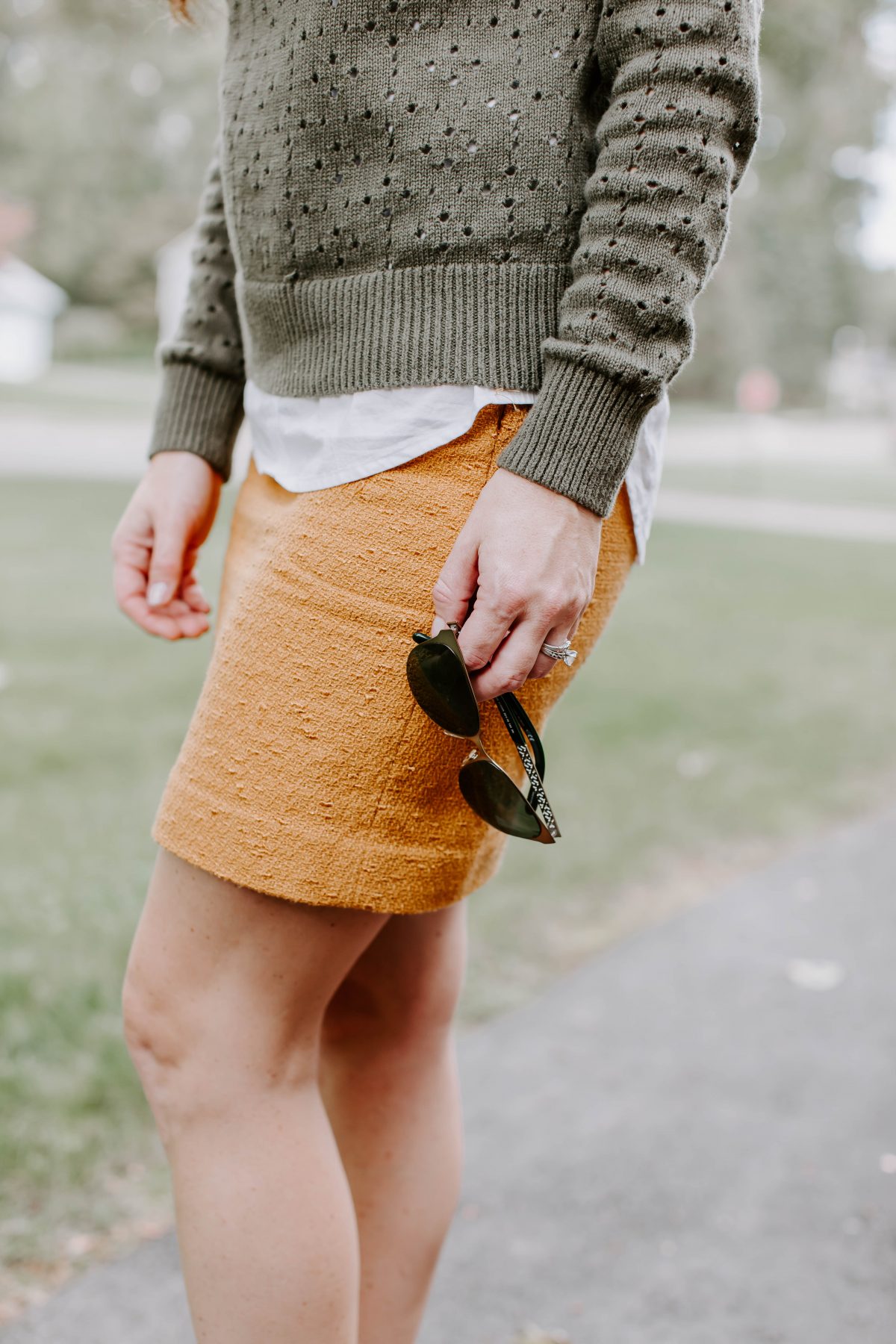 Detail shot of woman holding sunglasses while wearing green sweater and mustard skirt 