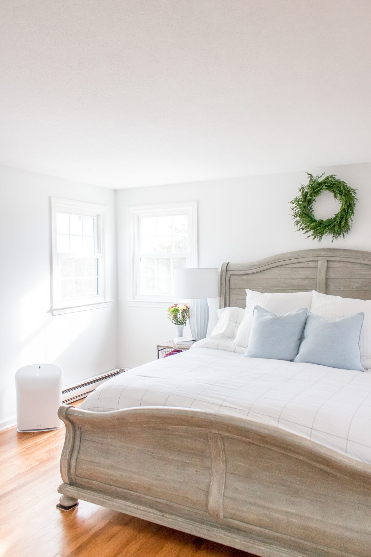 Bright White Bedroom with Gray Headboard and Green Boxwood Wreath on the Wall above the Bed and Rabbit Air Purifier near the wall in the room
