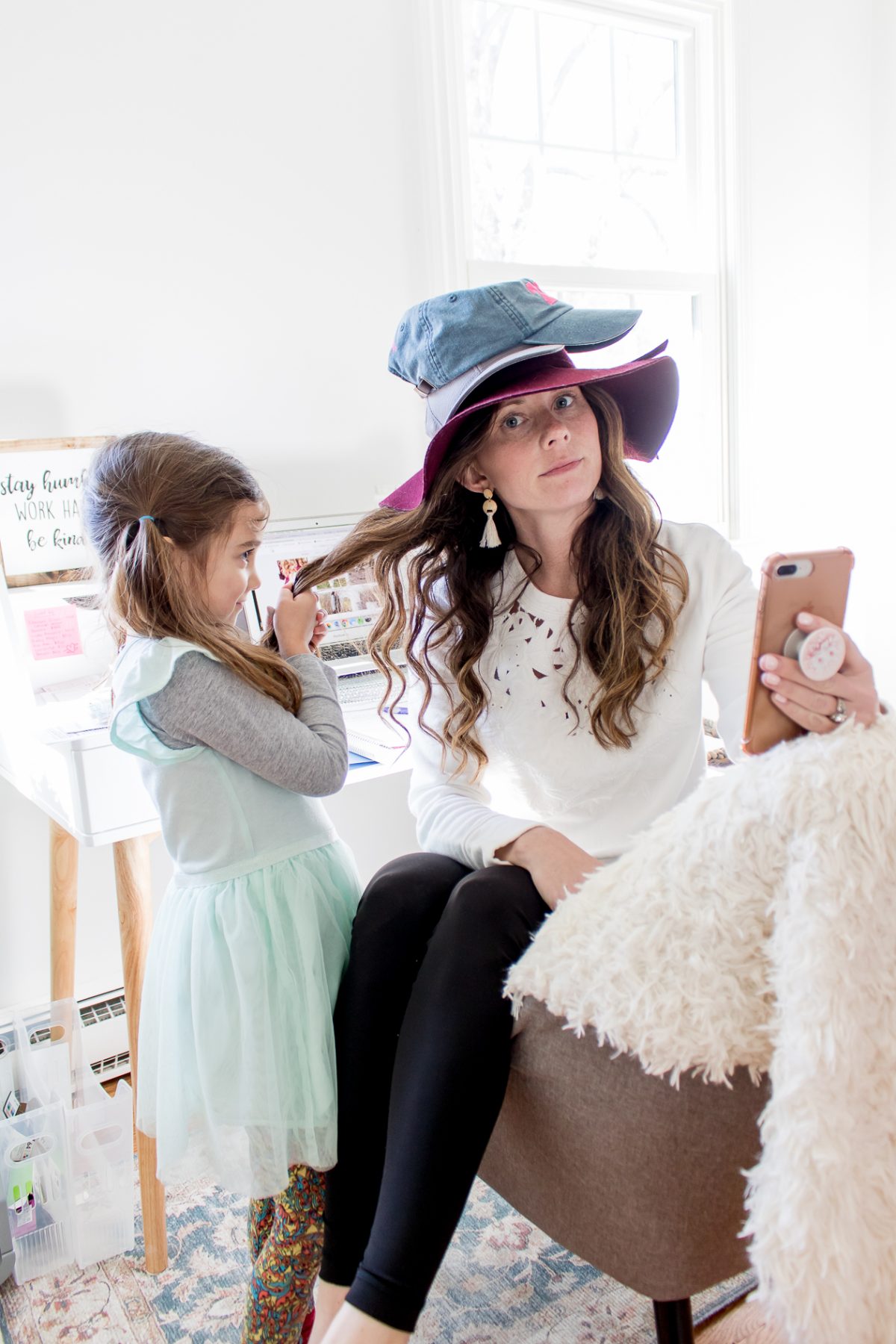 mom wearing three hats looking at the camera and holding a phone while her daughter pulls her hair 
